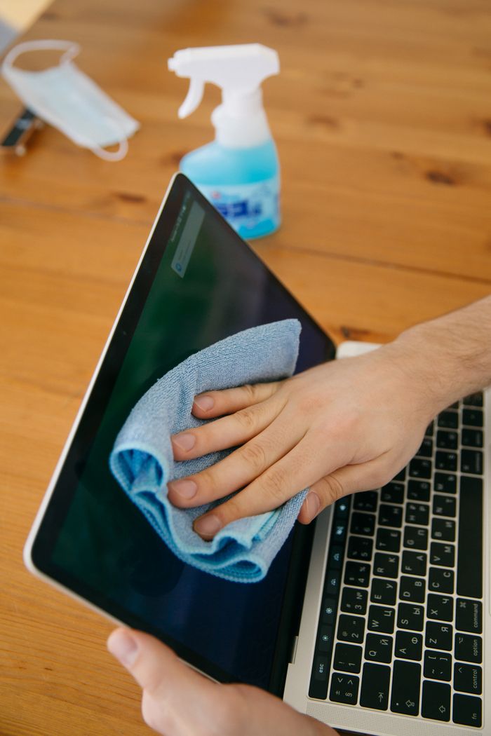 portfolio-03 A person wipes a laptop screen using a blue cloth and spray bottle on a wooden table for hygiene.