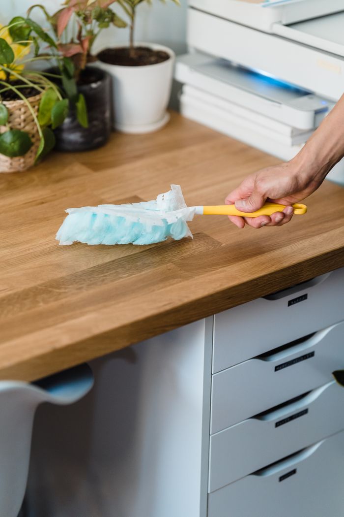 portfolio-04 A hand using a feather duster to clean a wooden desk, surrounded by plants and office items.